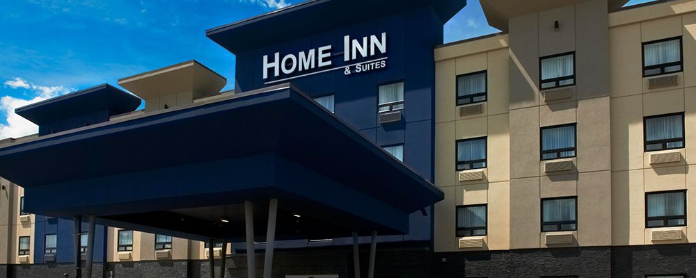 An upfront view of the flat roof navy blue portico that provides coverage over the entrance to the d3h Home Inn & Suites Saskatoon South. The corporate name, in bold white typeset is displayed as signage high above the hotel entrance and portico.