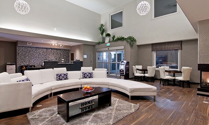 The hotel lobby of d3h HOME INN & SUITES BY d3h™ REGINA AIRPORT features hardwood flooring and a semi-circular white sectional sofa, encircling a solid dark wood coffee table placed over an area rug.  The charcoal gray check-in counter is contrasted against a white and dark patterned accent back wall, while the ATM and two square tables, complemented by white upholstered Parsons chairs, are set at the periphery of the lobby.