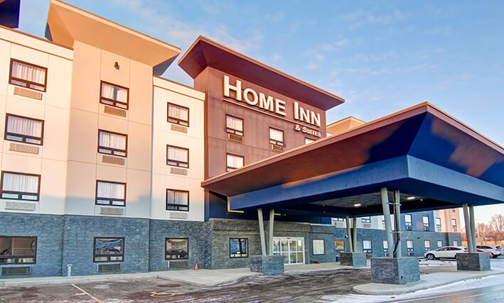 The exterior parking lot view of the four storey building of d3h HOME INN & SUITES BY d3h™ SASKATOON SOUTH  on a snowy day in Saskatchewan.  A large concrete portico with four steel and concrete posts provide coverage over the back entrance to the hotel, while several parked cars can be seen in the distance.
