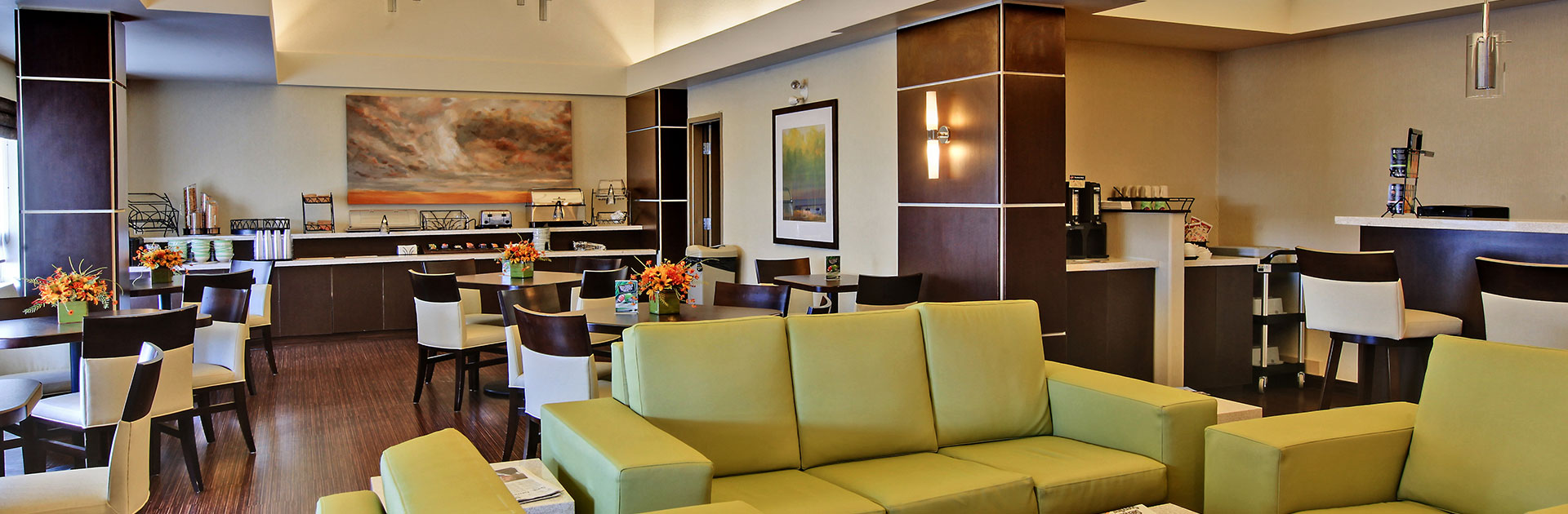 The HomeEssentials breakfast area of d3h Homesuites Regina East is equipped with a long two tier breakfast counter laden with plastic cups, stacked bowls, condiments and stainless steel appliances such as a toaster and countertop food warmer.  Square eating tables encircled by white leather upholstered dining chairs are placed throughout the eating space. The lounge area is furnished with a light chartreuse leather sofa and armchair set.