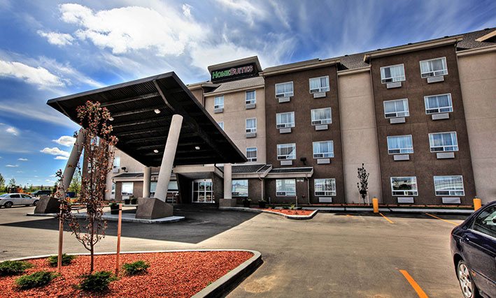 The d3h HOMESUITES BY d3h™ REGINA East hotel is a four storey building painted in two tone chocolate brown and beige.  The HomeSuites corporate name is presented in red and green lettering signage positioned at the summit of the hotel.  The parking lot is relatively empty with two vehicles parked nearby the tilted rectangular portico supported with concrete columns.