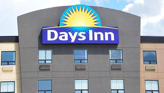 A large scale sign of the Days Inn logo is placed high atop the roof of the graphite-gray exterior of a Days Inn property.  The Days Inn corporate name is presented in bold white type, with a yellow sunrise icon contrasted against a blue background.