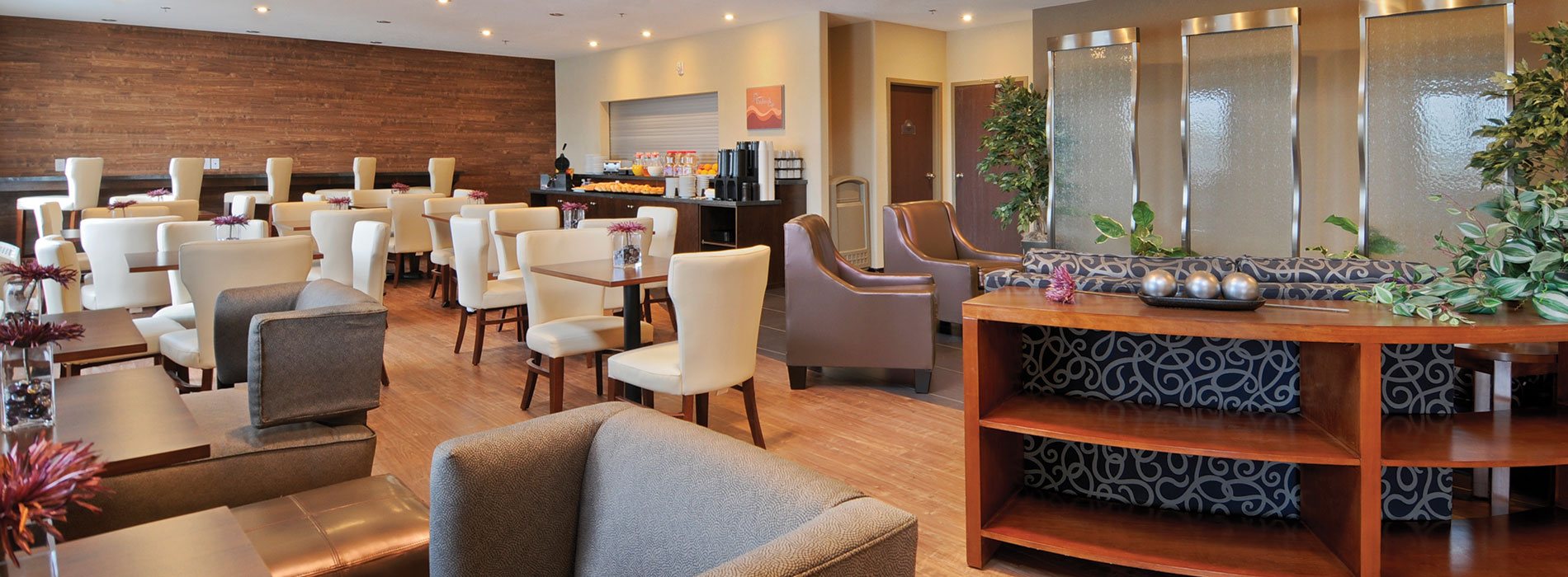 An overview of the Daybreak Café and the lounging space at d3h Days Inn Regina Airport West.  The lounge area is furnished with a swirl patterned navy blue sofa, two vinyl latte color armchairs, a sofa console table and several placements of potted green leaf plants.  The dining space features a fully stocked chocolate brown breakfast counter, a tawny brown wood paneled accent wall with built in breakfast bar and square eating tables paired with white upholstered dining chairs.