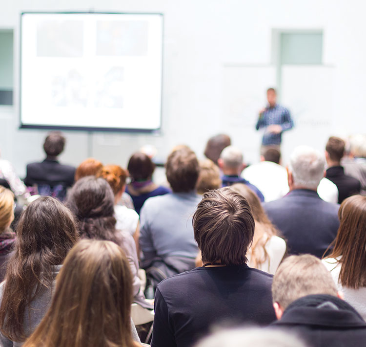 A group of men and woman in a lecture hall, all heads turned toward a speaker up front in the distance giving a presentation using a projector screen.