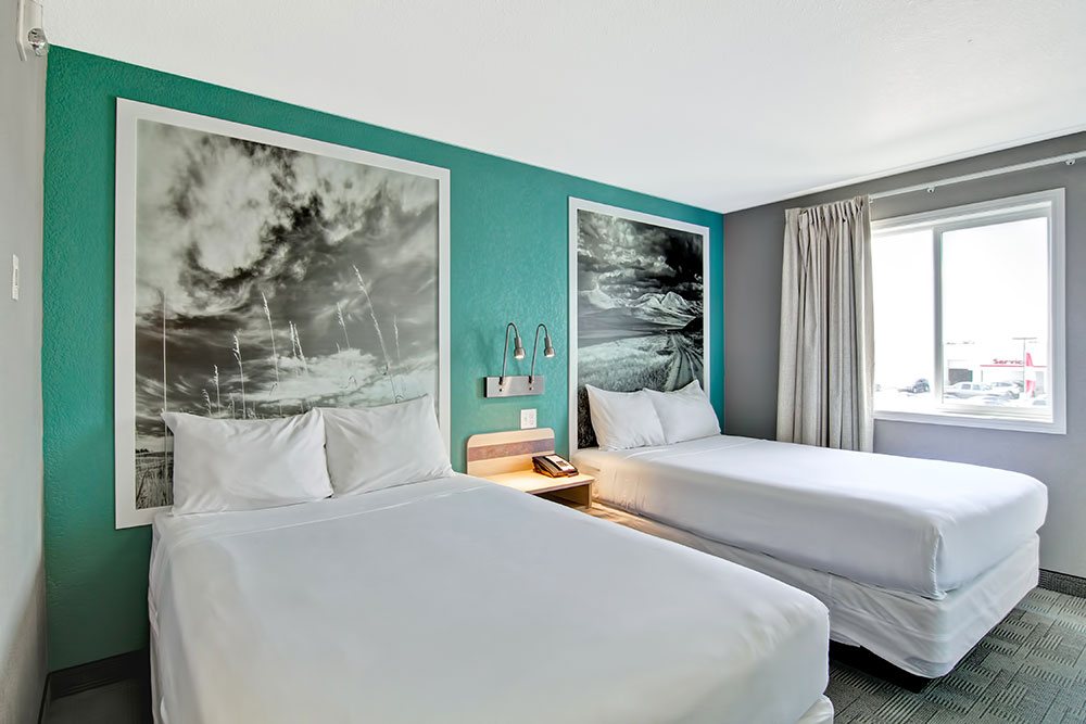 The Family Room suite at Home Inn Express in Medicine Hat features a tri-color interior: turquoise green, white and seal gray painted walls with large white framed black and white photos mounted against the walls.  Two wall mounted metal gooseneck lamps are placed between two beds dressed completely in white linen.
