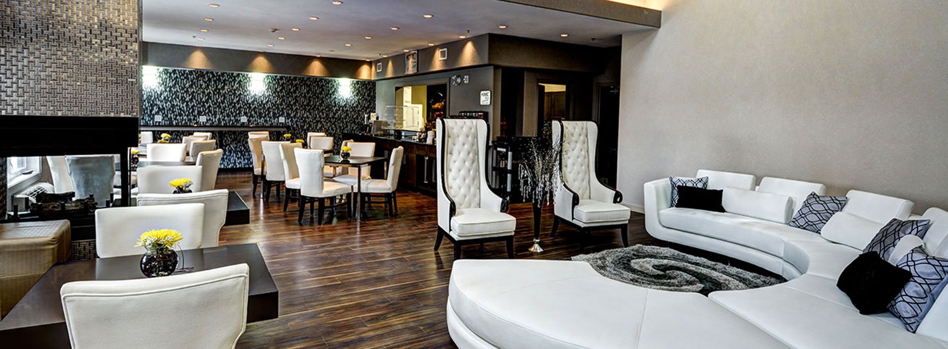 Pot lights and natural lighting provide an elegant ambience to the sitting and eating areas of d3h Home Inn & Suites Swift Current, featuring a glass enclosed fireplace with a shiny silver metallic surround in the corner of the room.  A white circular sectional sofa matched with white tufted high-backed, black-trim arm chairs and area rug define the lounging space.  Dark wood square tables and high end white Parsons chairs are placed throughout the eating space.