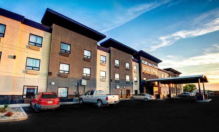 A small view of the back parking lot of d3h HOME INN & SUITES BY d3h™ YORKTON with several vehicles parked up to the three storey, two tone slate-gray and off-white building.  A semi-circle shaped portico hovers over the hotel entrance with the corporate name in presented in white lettered signage placed above.  A large orange banner inscribed in bold white lettering Now Open is draped beneath the corporate sign.