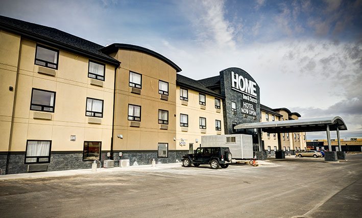 The view of the parking lot of the d3h HOME INN & SUITES BY d3h™ SWIFT CURRENT hotel on a clear day, with several vehicles parked next to the semi-circular metal portico covering over the entrance.  The corporate name (in large white lettering) is placed above the entrance, with a black banner advertising Hotel Now Open in bold white lettering typeset.