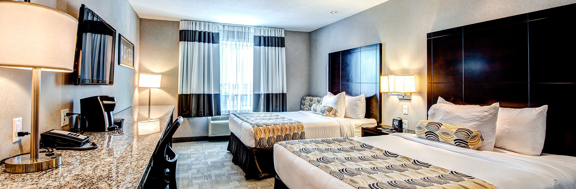 In the Dream Room at d3h Home Inn & Suites Yorkton, two queen sized beds (dressed in white and print linen) paired with tall dark headboards face an elongated marbled granite top work bench on the opposite side of the room.  A wall mounted flatscreen TV sits above the bench, on which sits a phone, coffeemaker and gold brass table lamp.