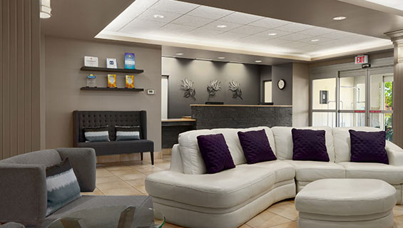 Plush white two sectional sofa with matching ottoman, a single gray chair with shortened arms and charcoal loveseat provide ample seating in the lobby of d3h Days Inn Edmonton South. The matching charcoal gray stone countertop of the check-in counter sits in the background with three floral metal wall ornaments against slate gray back walls.