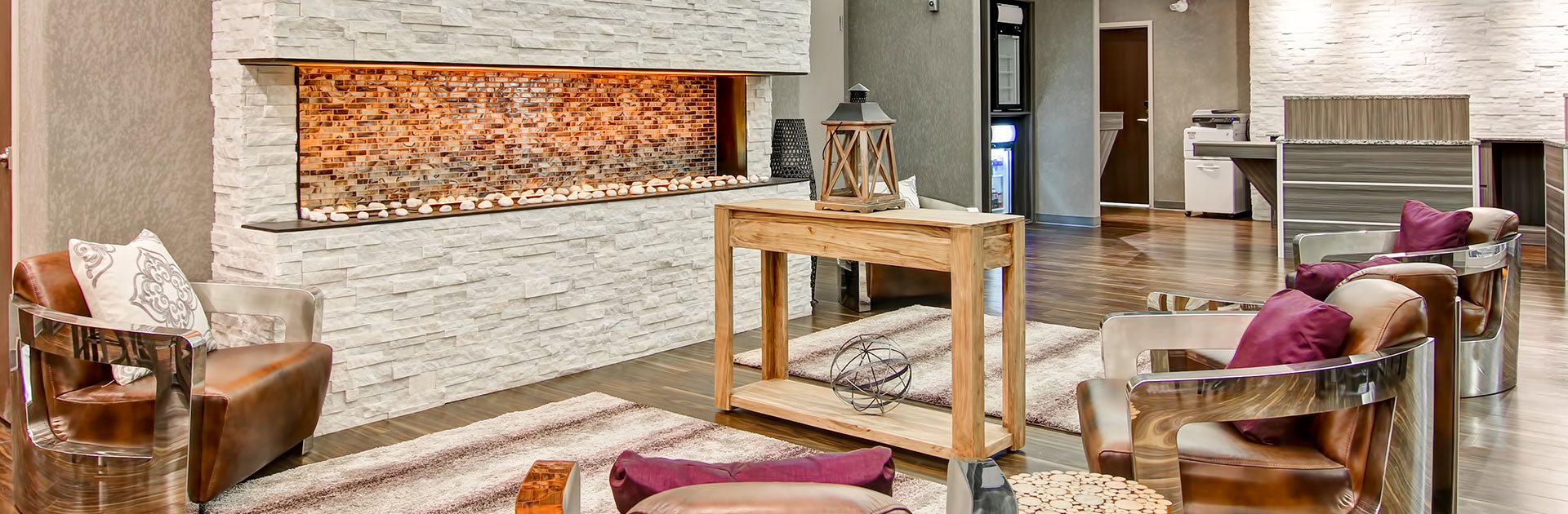 The centre of the lounge area at d3h Home Inn & Suites Saskatoon South showcases a towering white stacked limestone electric fireplace, with a recessed brick interior.  Leather club chairs, a rustic wood grain console table displaying carved wood and wrought iron art decor, and area rugs add to the coziness and comfort of the lounge.