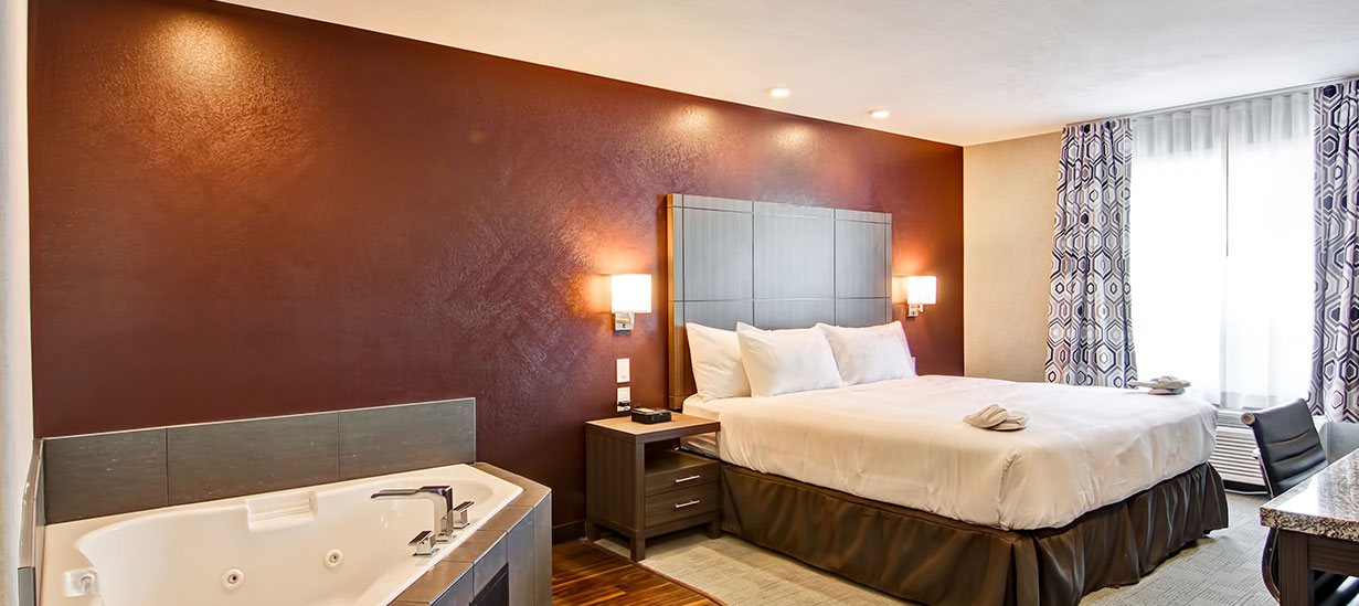 The Jacuzzi suite at d3h Home Inn & Suites Saskatoon South features a gray tiled soaker hot tub in the corner of the room.  A king sized bed and light mocha wood paneled headboard and matching bedside table add to the decor of the spacious suite, while the sienna brown accent wall is contrasted against white adjacent walls and ceiling.