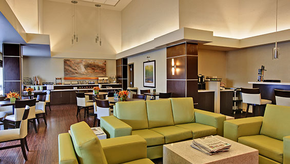 Dark hardwood floors run throughout the eating and lounging space of the d3h HomeSuites Regina East, with a dark wood two tier breakfast counter displaying stainless steel appliances, table and eating ware.  Square dining tables with chairs are placed next to the lounge area furnished with chartreuse green sofa and armchair sets and a solid white cube coffeetable.  A coffee kiosk is discreetly tucked away in a corner behind a wood paneled wall.