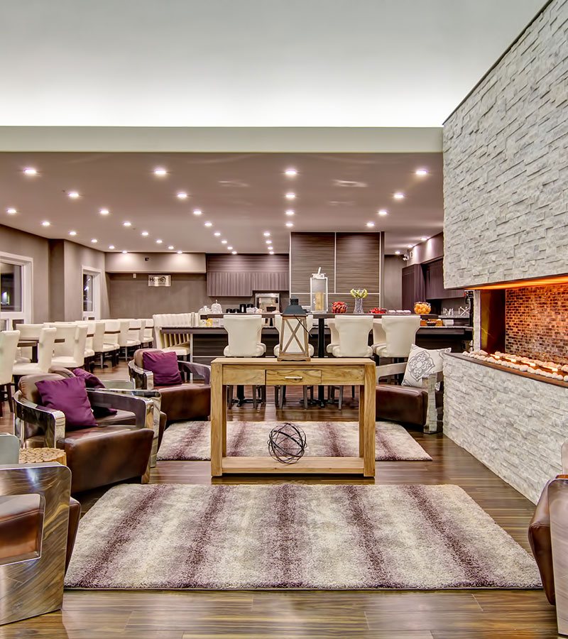 The HomeEssentials breakfast bar at d3h Home Inn & Suites Saskatoon South features an eat-in counter (stocked with fruit, counter appliances and tableware), eating tables and dining bench set amidst a sea of white upholstered Parsons chairs.  The adjacent lounging area showcases a floor to ceiling birch limestone electric fireplace surrounded by leather and metal clubchairs.