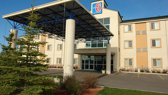 The back entrance to d3h Motel 6 Red Deer, covered over with a rectangular metal portico with concrete post supports.  At the top of the three storey hotel, the corporate logo (a red and bold number 6 against a backdrop of blue), used as signage, overlooks an empty parking lot and Jack pine trees planted in a landscaped area on the property.