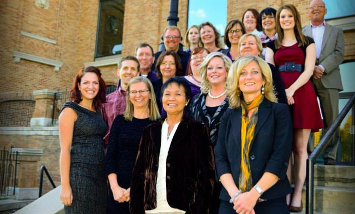 Smiling male and female employees of d3h hotels stand before the steps of a faded brick building for a photo shoot.